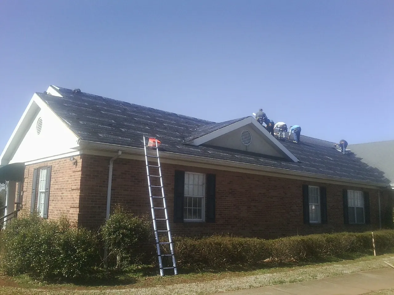Crew tearing shingles off a brick ranch roof with a ladder set — Emergency Roof Repair work by our crew in East Branch
