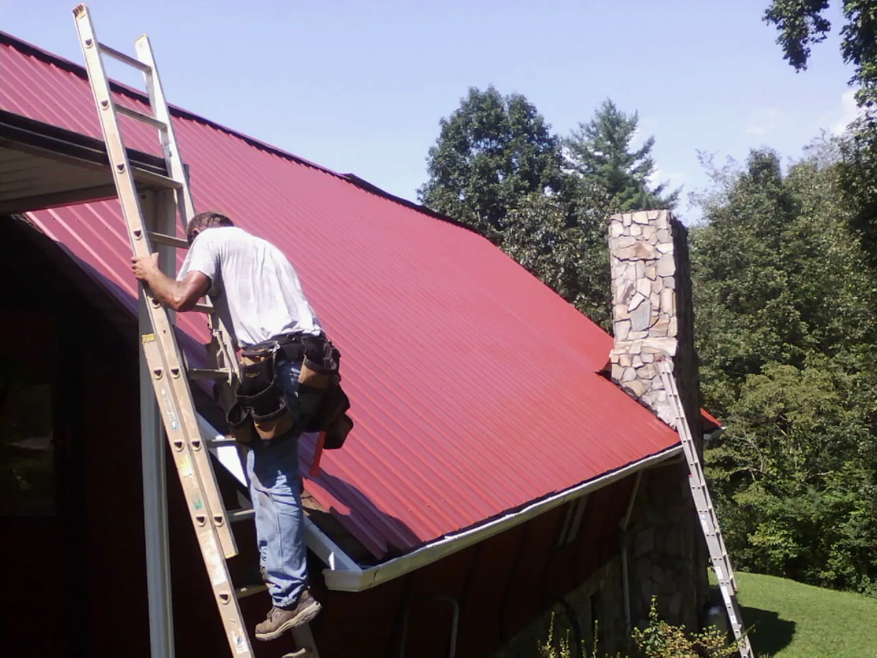 Roofer on a ladder beside a red metal roof and stone chimney — Soffit & Fascia Repair by Roofing Brothers in East Branch